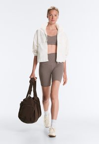 Femme marchant vers l'avant portant un crop top à carreaux et un short, une veste blanche, des chaussettes blanches, des baskets, portant un grand sac marron foncé.