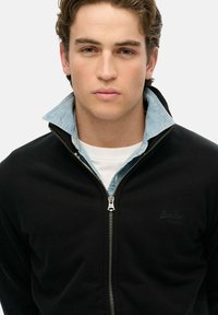 Giacca nera con zip e colletto alto, indossata sopra una camicia di jeans azzurro chiaro. Presenta una zip frontale e un dettaglio del logo sul petto.