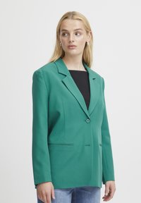 Blazer verde con un solo botón, solapas dentadas y dos bolsillos frontales. La tela tiene una textura suave y un diseño ajustado.