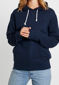 Person klädd i en marinblå hoodie med vita dragsnören och ljusblå jeans, händerna knäppta framför sig, mot en enkel bakgrund.
