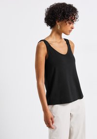 Top negro sin mangas con escote redondo y detalle de hombro fruncido, confeccionado en una tela suave para un ajuste relajado. Combinado con pantalones beige claros.