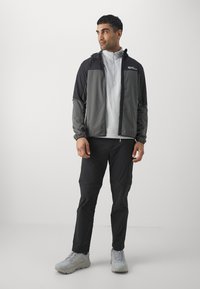 Mannelijke sportoutfit met een grijze en zwarte ritsjack, lichtgrijze basislaag en zwarte broek, met gestructureerde stof en ritsaccenten.