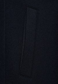 Czarna wełniana tkanina płaszcza z subtelną fakturą, z prostokątną kieszonką wszytą w projekt.