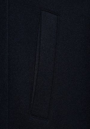 Czarna wełniana tkanina płaszcza z subtelną fakturą, z prostokątną kieszonką wszytą w projekt.