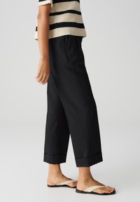 Pantalons noirs à jambes larges avec ourlet retroussé, associés à des sandales beiges à brides croisées. L'ensemble présente une coupe décontractée et un design minimaliste.