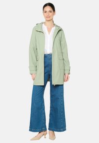 Lichtgroene zip-up jas met capuchon, voorzien van knoopzakken en een relaxte pasvorm. Gecombineerd met wijde blauwe jeans en beige hakken.