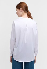 Camicia bianca a maniche lunghe con texture liscia, collo rotondo e dettaglio di piega sul retro, abbinata a jeans blu a vita alta.