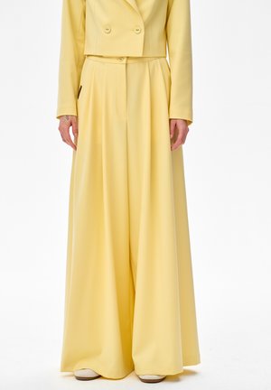 Kobieta ubrana w żółte spodnie o szerokich nogawkach i wysokim stanie, zestawione z dopasowaną, skróconą marynarką dwurzędową, stoi z rozluźnionymi ramionami.