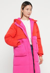 Giacca imbottita bicolore con maniche rosse e corpo rosa, dotata di cappuccio e tasche, realizzata in materiale leggero con una trama liscia.