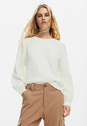 Maglione bianco con maniche a treccia, polsini a coste e scollatura rotonda, abbinato a pantaloni beige chiaro con chiusura a bottoni.