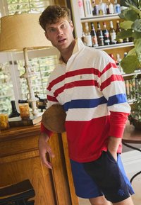 Punases ja valges triibuline ragbi särk sinise triibuga, pikkade varrukatega ja kraega; koos siniste lühikeste pükstega. Hoides pruuni jalgpalli.