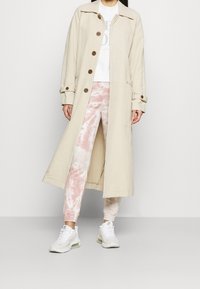 Beżowy długi płaszcz z guzikami, noszony na białej grafitowej bluzie i różowych spodniach dresowych w stylu tie-dye. Do tego białe buty sportowe.