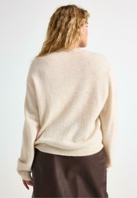 Beige strikket sweater med en afslappet pasform og lange ærmer, der har en blød tekstur og ribbet kant. Båret over en mørkebrun læderskirt.