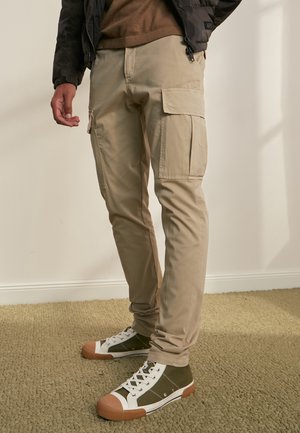 Man die beige cargobroek en groene hoge sneakers draagt, staande op een beige tapijt in een goed verlichte kamer.