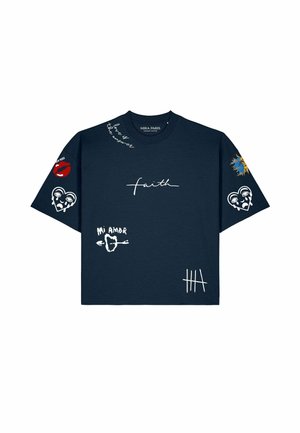 Man draagt een oversized marineblauw T-shirt met witte en rode grafische prints, lichtblauwe spijkerbroek en grote zonnebril met schildpadrand.