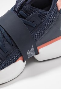 Basket Reebok bleu foncé avec tige en maille, large sangle sur les lacets, semelle blanche et accent rose clair entre les couches de la semelle.