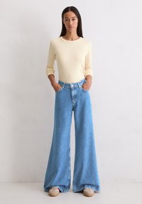 Vrouw die tegen een witte muur staat, gekleed in een crèmekleurig shirt met lange mouwen, wijde blauwe jeans en beige sneakers met rode accenten.