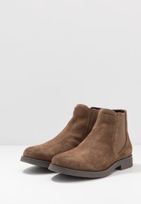 Botas de tobillo de ante marrón con una textura suave, punta redondeada, paneles laterales elásticos y un tacón de goma bajo para estabilidad y comodidad.