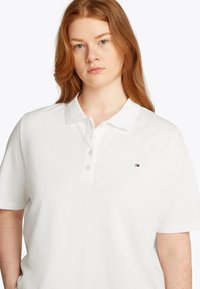White polo shirt with short sleeves, three button placket, collar, and a small colored logo on the left chest. Smooth fabric texture.