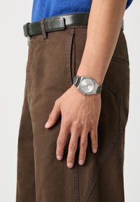 Montre en acier inoxydable argenté avec un cadran rond et un design minimaliste, portée sur une main, associée à un pantalon marron et un haut bleu.