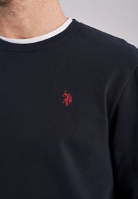 Marinblå bomulls sweatshirt med rund halsringning, prydd med en diskret röd broderad logotyp på bröstet och en vit detalj vid innerkragen.