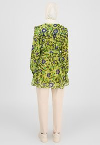 Green floral print blouse with ruffled collar and long sleeves, cinched waist, sheer material, paired with beige pants and sneakers.