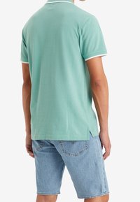 Polo vert clair avec des accents blancs, manches courtes et fentes sur les côtés ; associé à un short en denim bleu pour un look décontracté.
