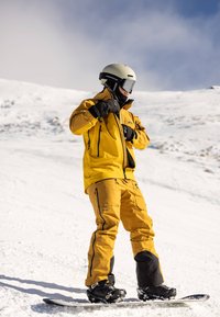 Żółta kurtka i spodnie narciarskie z czarnymi akcentami, połączone z czarnymi butami snowboardowymi, stojące na śniegu z deską snowboardową pod spodem.