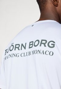 Λευκή κοντομάνικη μπλούζα με μαύρο κείμενο που γράφει "BJÖRN BORG TRAINING CLUB MONACO" εμφανώς στην πλάτη. Λείο υφασμάτινο υλικό.