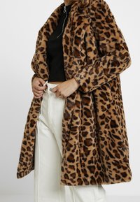 Cappotto in pelliccia sintetica con stampa leopardata, colletto, apertura frontale e maniche lunghe. Indossato sopra un top nero e pantaloni bianchi. Texture morbida.