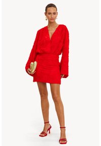 Vestido rojo texturizado con mangas largas y un escote en V profundo. La falda es ajustada. Combinado con stilettos rojos y un clutch adornado con dorado.