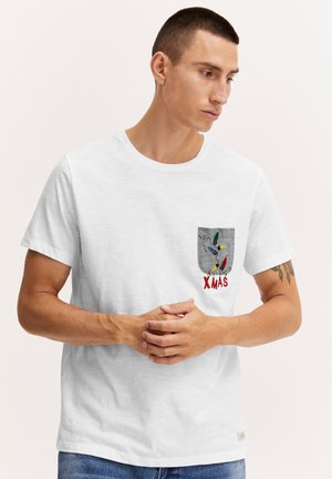 Mężczyzna w białej koszulce z szarą kieszenią na piersi ozdobioną kolorowymi światełkami świątecznymi i napisem "XMAS", stoi ze złączonymi rękami.