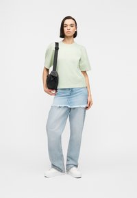 Γυναίκα στέκεται φορώντας τοπ με φουσκωτά μανίκια σε ανοιχτό πράσινο, ανοιχτά μπλε τζιν με επικάλυψη από μίνι φούστα με ξεφτισμένες άκρες, λευκά αθλητικά παπούτσια και μαύρη τσάντα ώμου.