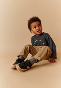 Grå långärmad skjorta med ett guld-elefantmotiv, beige byxor och svarta sneakers med en beige sula. Randiga grå strumpor är synliga.