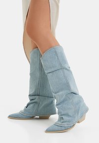 Helle blaue Jeans-Kniestrumpfstiefel mit einem lässigen Design, spitzer Zehe und einem niedrigen, gestapelten Absatz. Nähte sind entlang der Seiten sichtbar.
