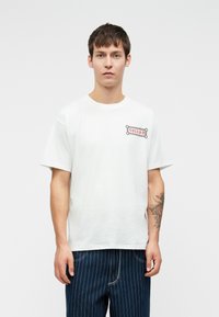 Witte katoenen t-shirt met korte mouwen, voorzien van een rood en grijs geborduurd logo op de linkerkant van de borst, gecombineerd met donkerblauwe gestreepte broek.