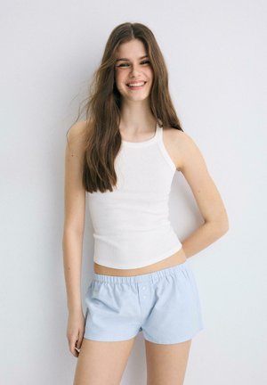 Giovane donna con lunghi capelli castani che sorride, indossa una canotta bianca senza maniche e pantaloncini azzurri, in piedi contro uno sfondo chiaro e uniforme.