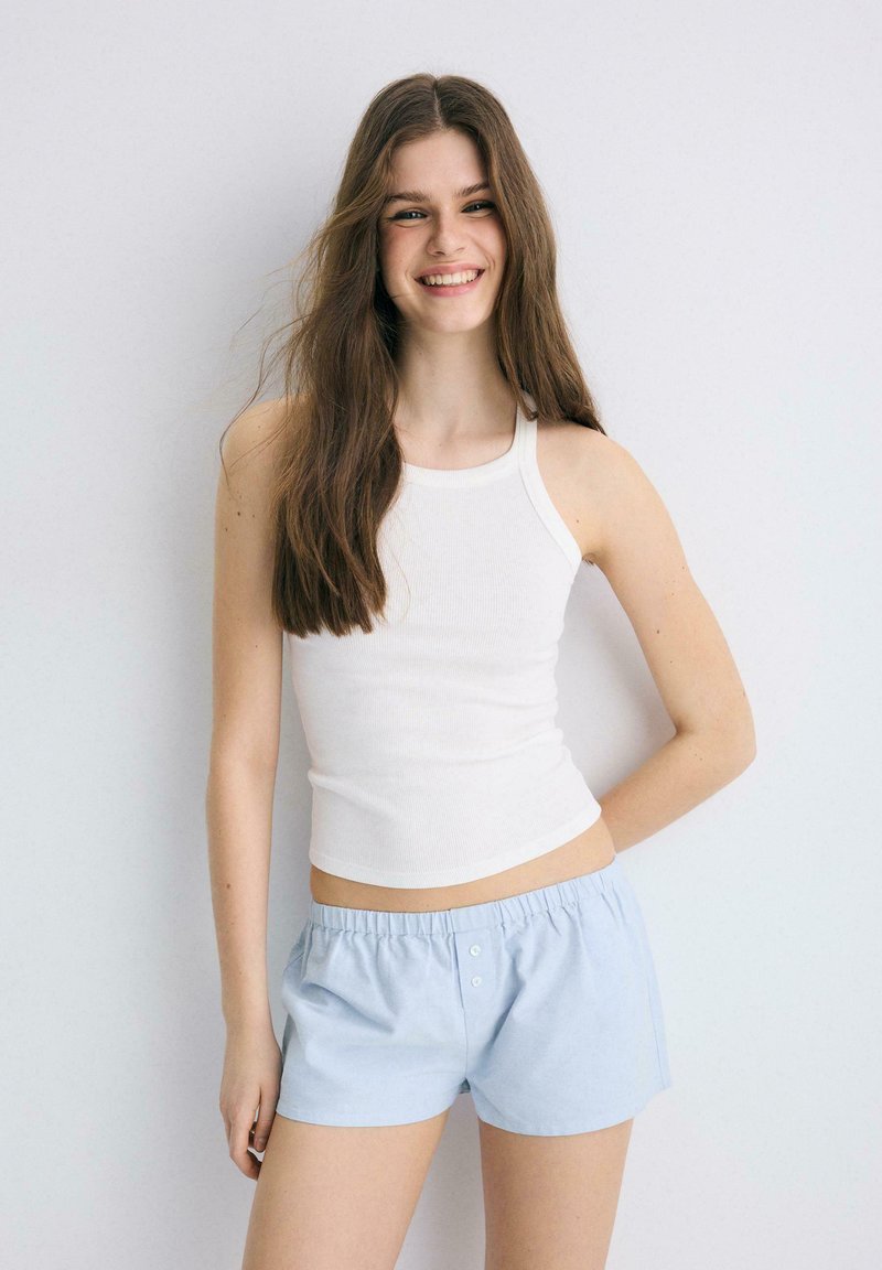 Jeune femme aux longs cheveux bruns souriante, portant un haut blanc sans manches et un short bleu clair, debout devant un fond clair uni.