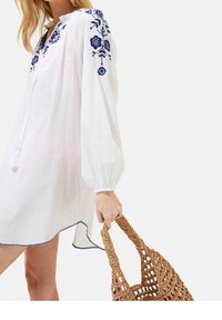 Abito bianco in lino con ricami floreali blu, maniche arricciate e dettaglio con nappine. Indossato con una borsa di paglia intrecciata, caratterizzata da un design testurizzato.