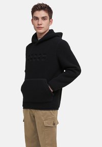 Haut en polaire noir avec une poche kangourou et un détail de texte en relief sur le devant, accompagné d'un pantalon cargo kaki avec des poches sur les côtés.