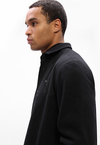 Fleece preto com gola, apresentando um bolso de peito com fecho éclair, tecido texturizado e um painel frontal suave.