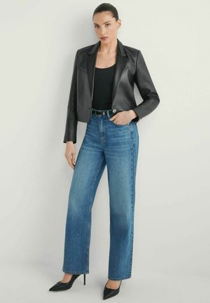 Kobieta ubrana w czarną, krótka skórzaną marynarkę na czarnym topie, niebieskie, wysokie proste jeansy i czarne szpilki z zaostrzonym noskiem, stojąca.