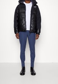 Svart puffjacka med blanka paneler, dragkedjestängning och justerbar huva. Matchad med blå skräddarsydda byxor och svarta ankelstövlar.