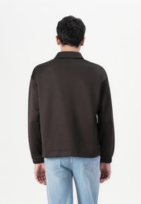 Pull-over marron à manches longues avec un col et des poignets élastiques, associé à un jean bleu clair. Texture lisse, coupe décontractée.