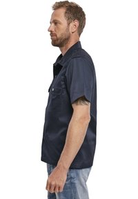 Camicia button-up a maniche corte di colore blu scuro, con due tasche sul petto, una texture liscia e una vestibilità rilassata, indossata con jeans azzurri.