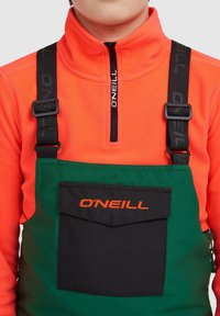 Overoles de tirantes para jóvenes en verde con un bolsillo negro, presentando tirantes ajustables negros. Debajo hay un forro polar naranja brillante con cremallera.