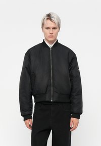 Chaqueta bomber negra con cremallera frontal, puños y dobladillo acanalados, y bolsillo lateral. Material suave y ligeramente brillante, corte corto.
