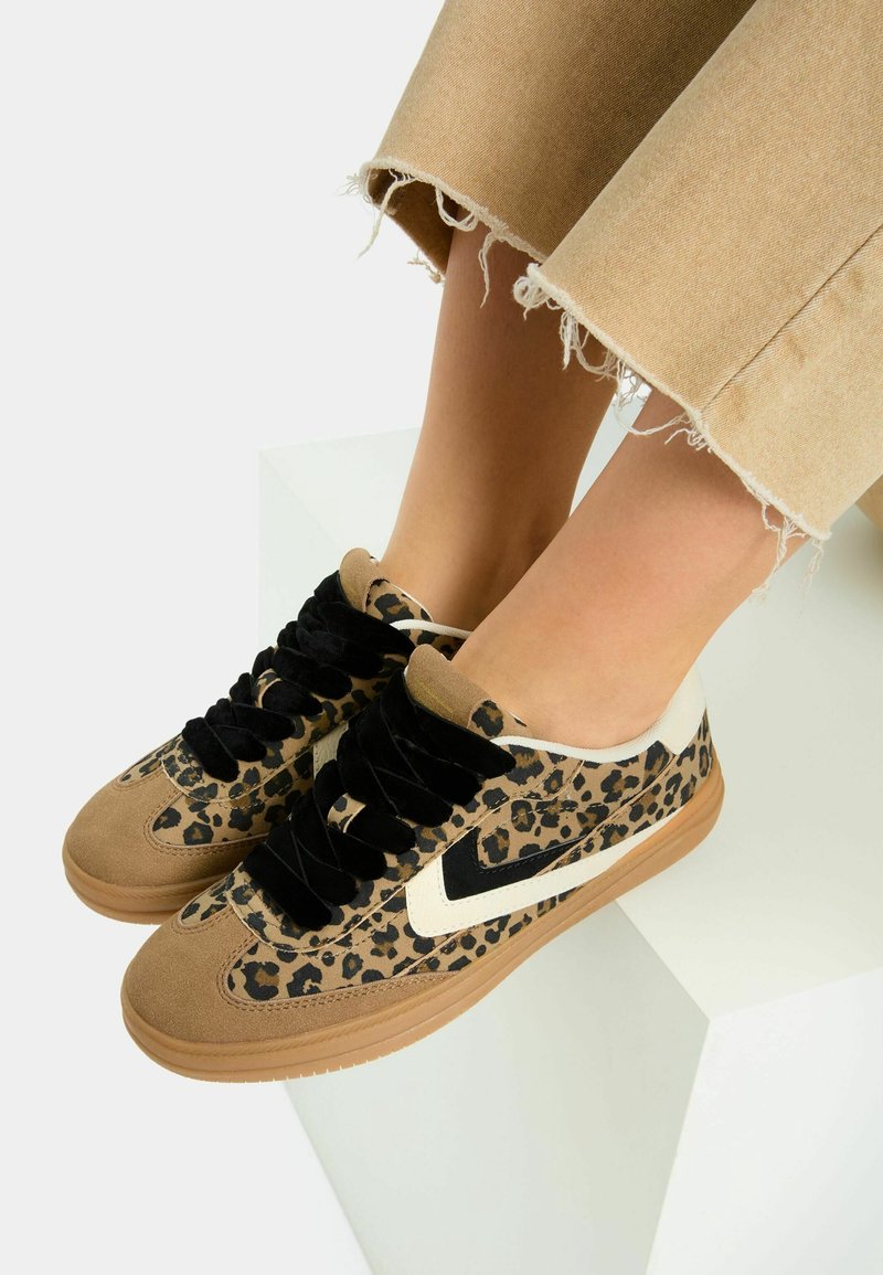 Pieds portant des baskets à imprimé léopard avec des lacets noirs et des détails en daim beige, associées à un pantalon beige court effrangé, reposant sur une surface blanche.
