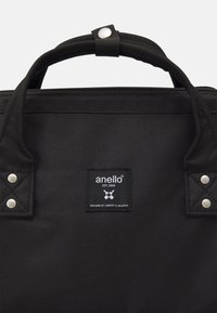 Czarna torba anello z teksturowanej tkaniny, z podwójnymi uchwytami u góry i prostokątnym kształtem. Zauważalne napy i przeszycia.