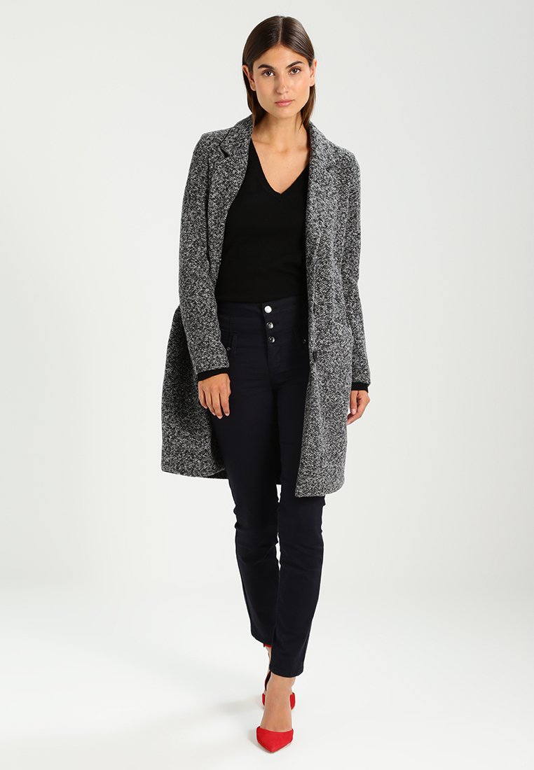 Übergroßer grauer und schwarzer gesprenkelter Mantel mit angedeutetem Kragen, getragen über einem schwarzen Oberteil, kombiniert mit dunklen Hosen und roten High Heels.